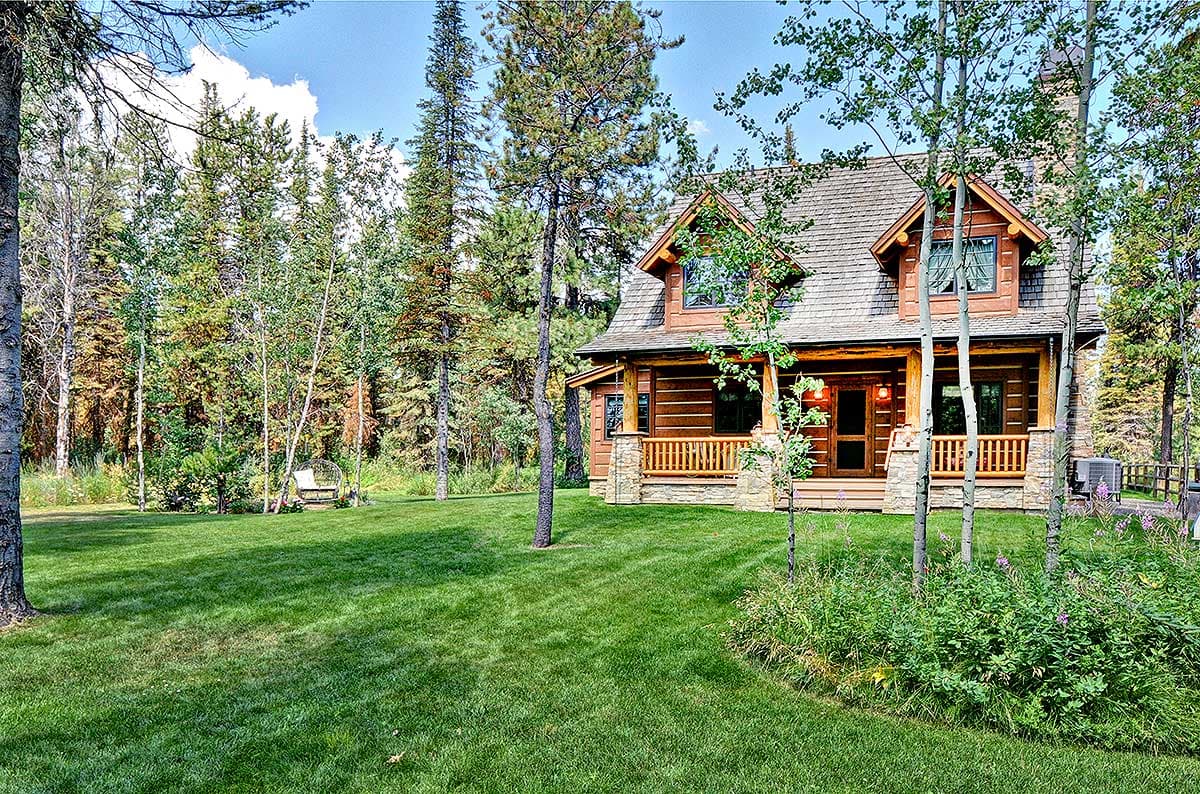 Log cabin exterior with a covered porch, stone pillars, multiple gables, and dormers, set among pine and aspen trees.