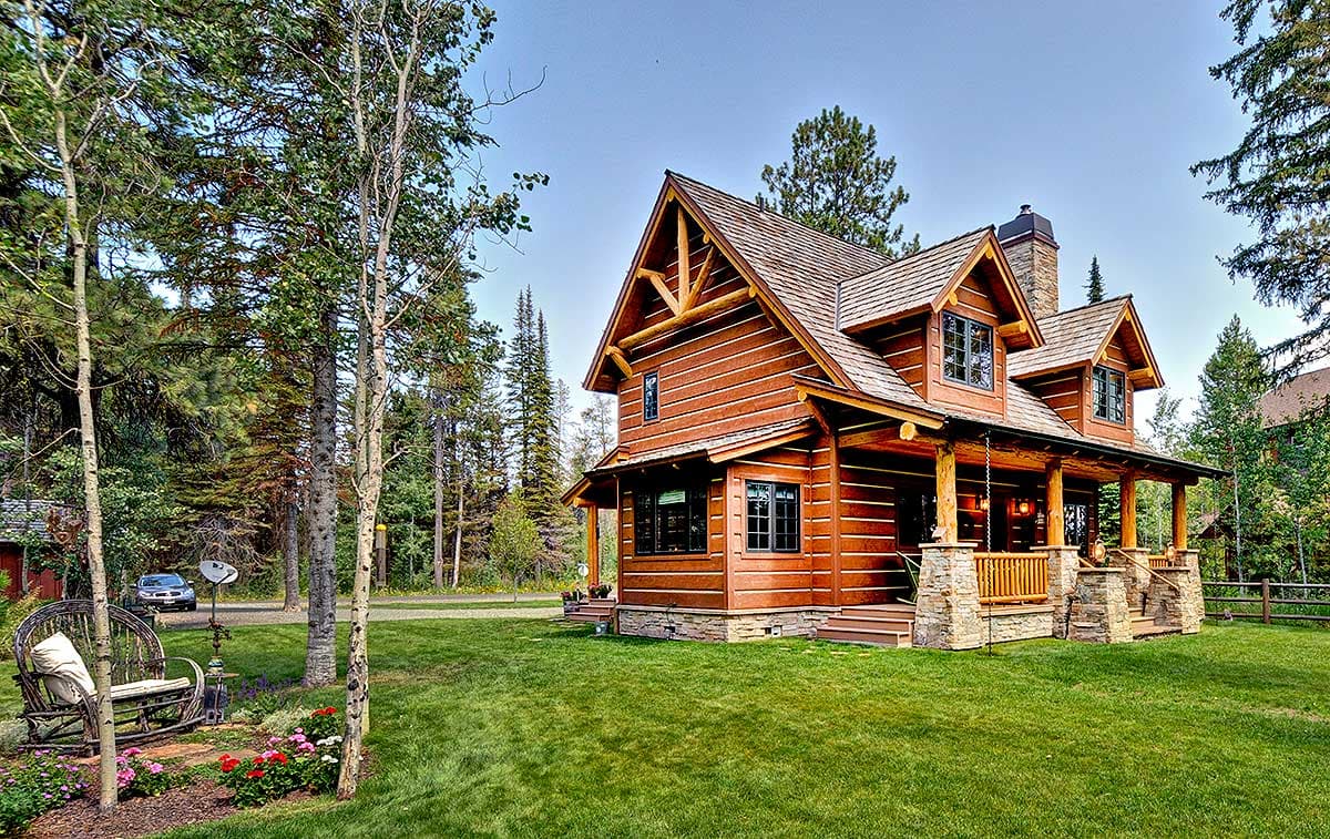 Log home exterior with wrap-around porch, stone foundation, and prominent gable dormers.