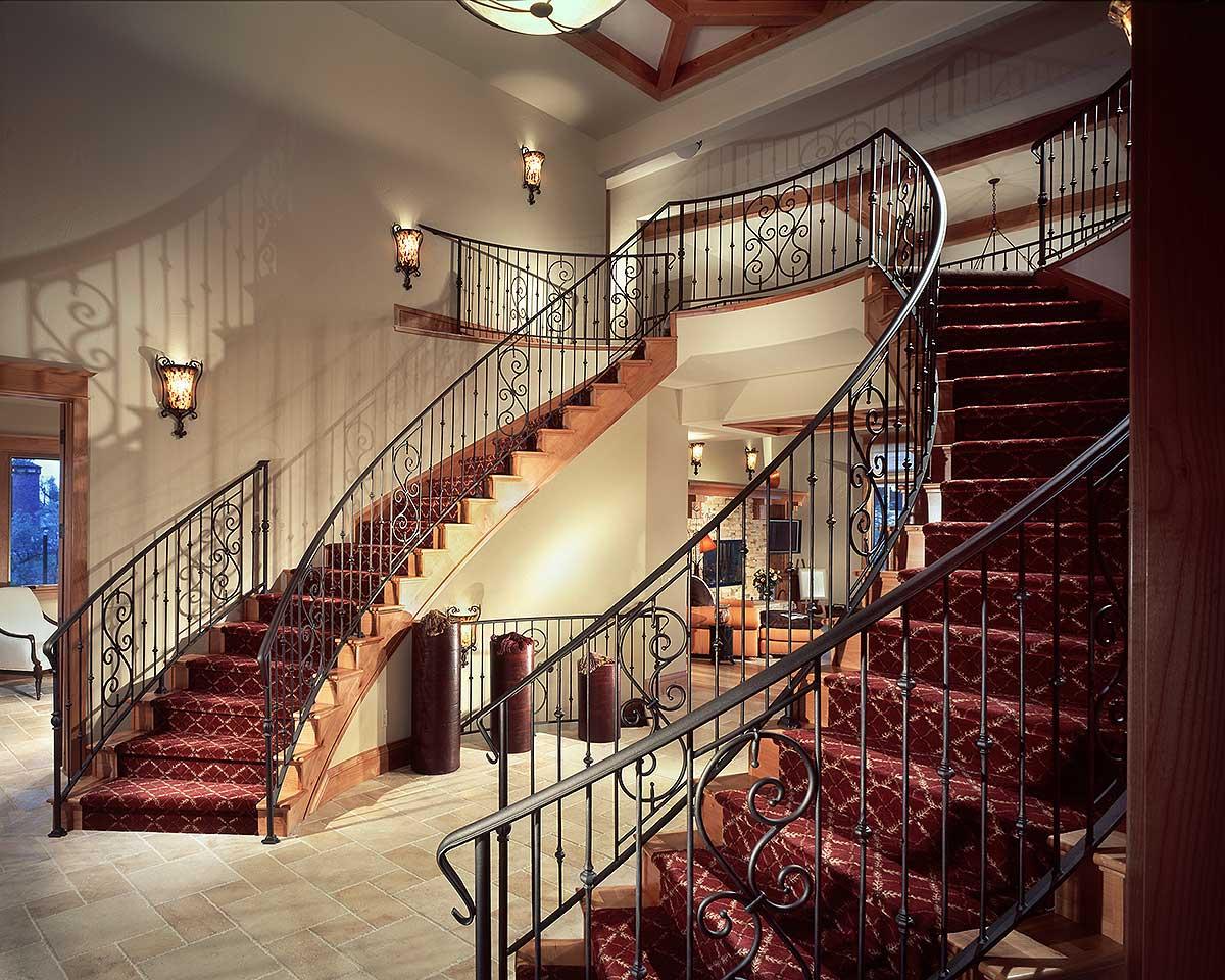 Grand foyer with dual curved staircases featuring ornate wrought iron railings and red patterned carpet runners.