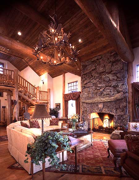 Living room with vaulted wood ceiling, large stone fireplace, antler chandelier, and open loft staircase.