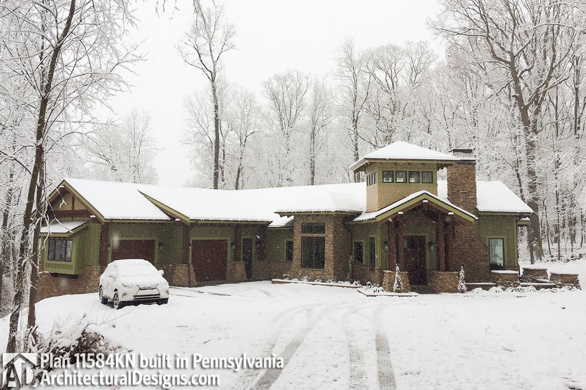 House plan exterior with green siding, stone accents, prominent chimney, covered entry, and three-car garage in a snowy setting.
