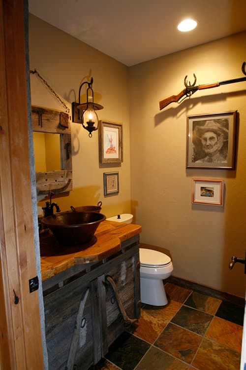 Bathroom with rustic vanity, copper sinks, toilet, and slate tile floor. Western decor includes a rifle and artwork.