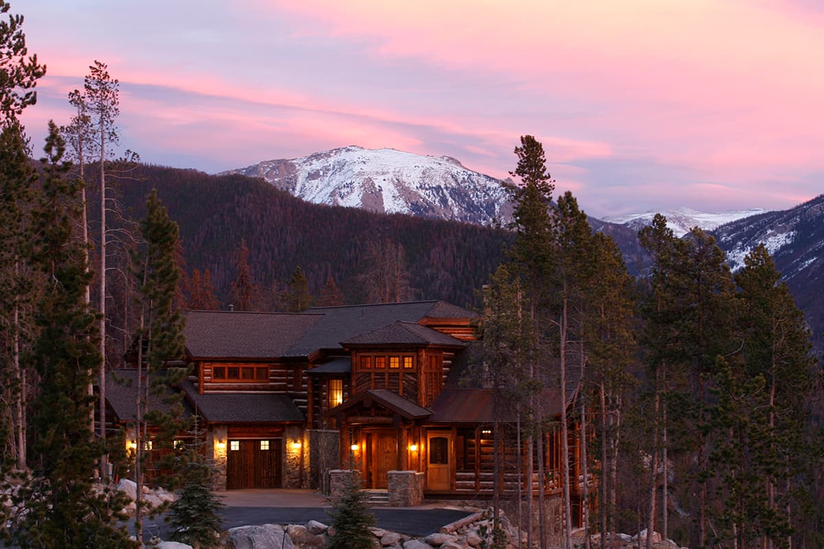 Log home exterior with multiple gables, stone accents, and a covered porch. Mountains and pine trees in background.