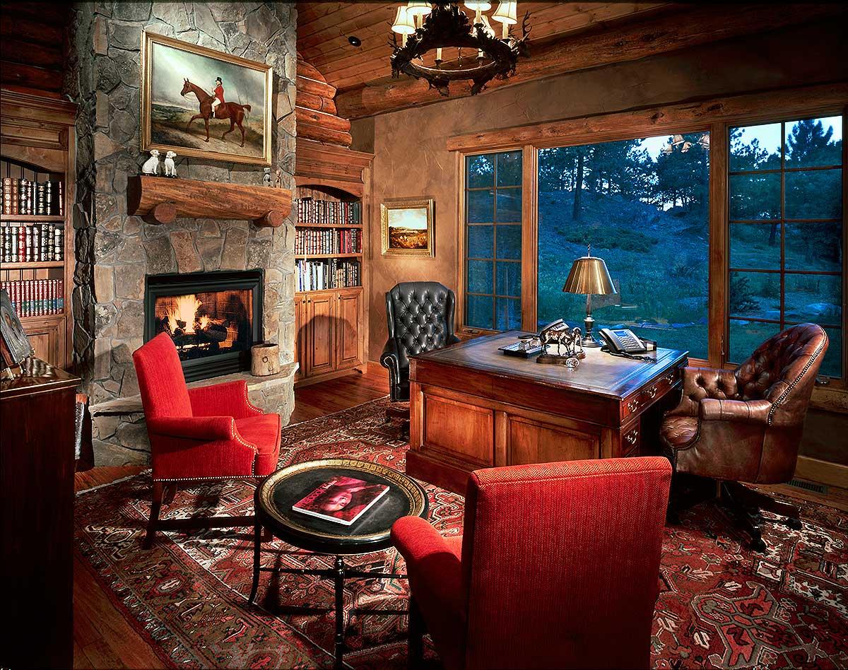Home office with stone fireplace, built-in bookshelves, large desk, leather chairs, and a scenic forest view through large windows.