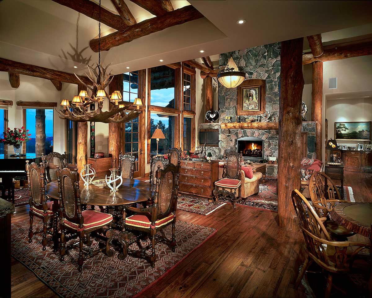 Rustic great room with log beams, stone fireplace, antler chandelier, and dining table.