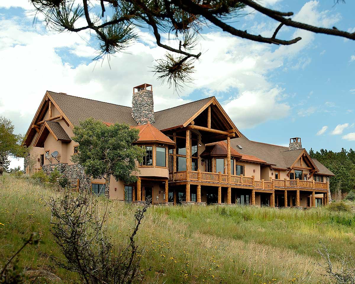 Log home exterior with prominent stone chimney, multi-level decks, and large exposed timber roof trusses.