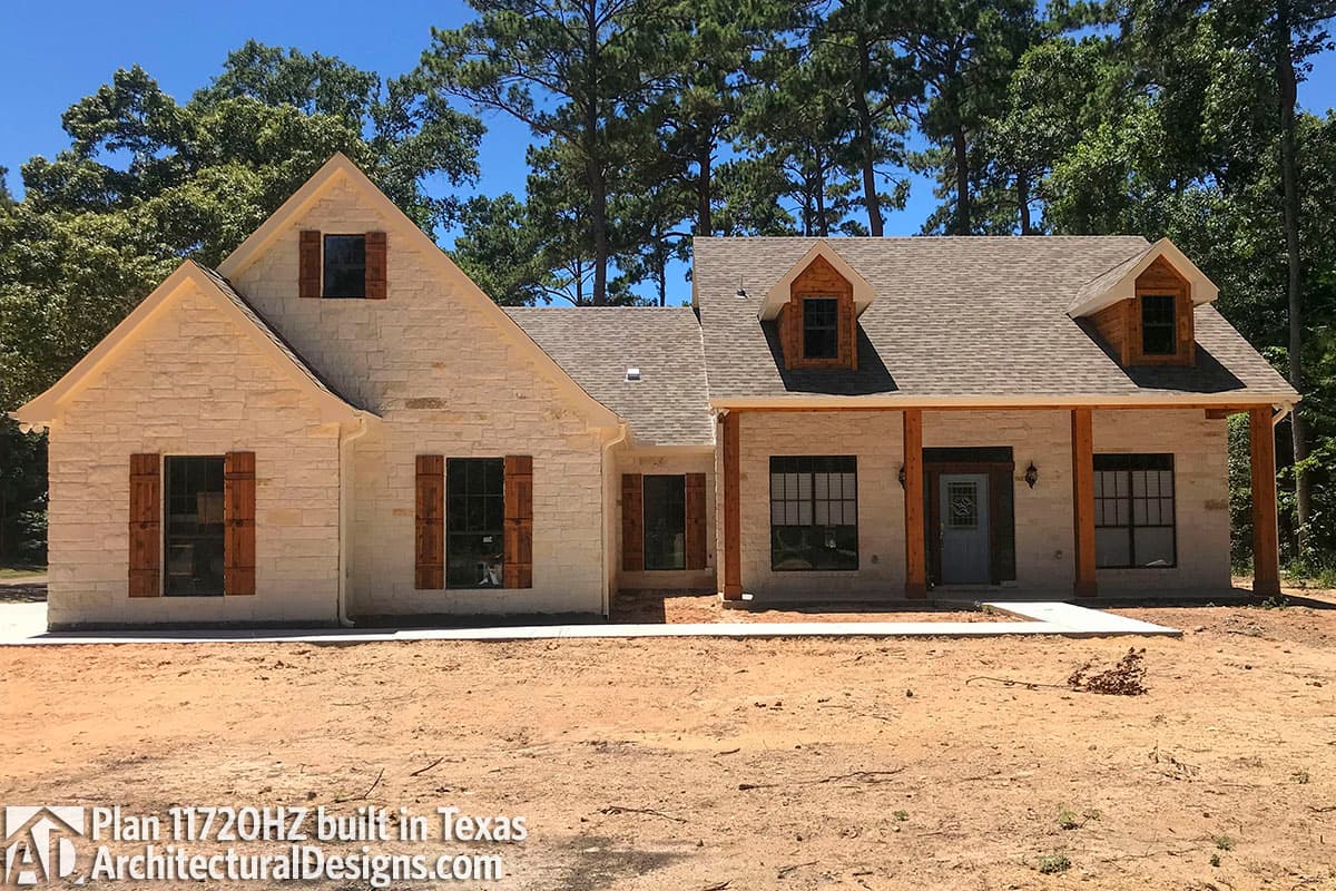 House plan exterior, one story, light stone veneer, gabled dormers, wood shutters, and covered porch with timber posts.