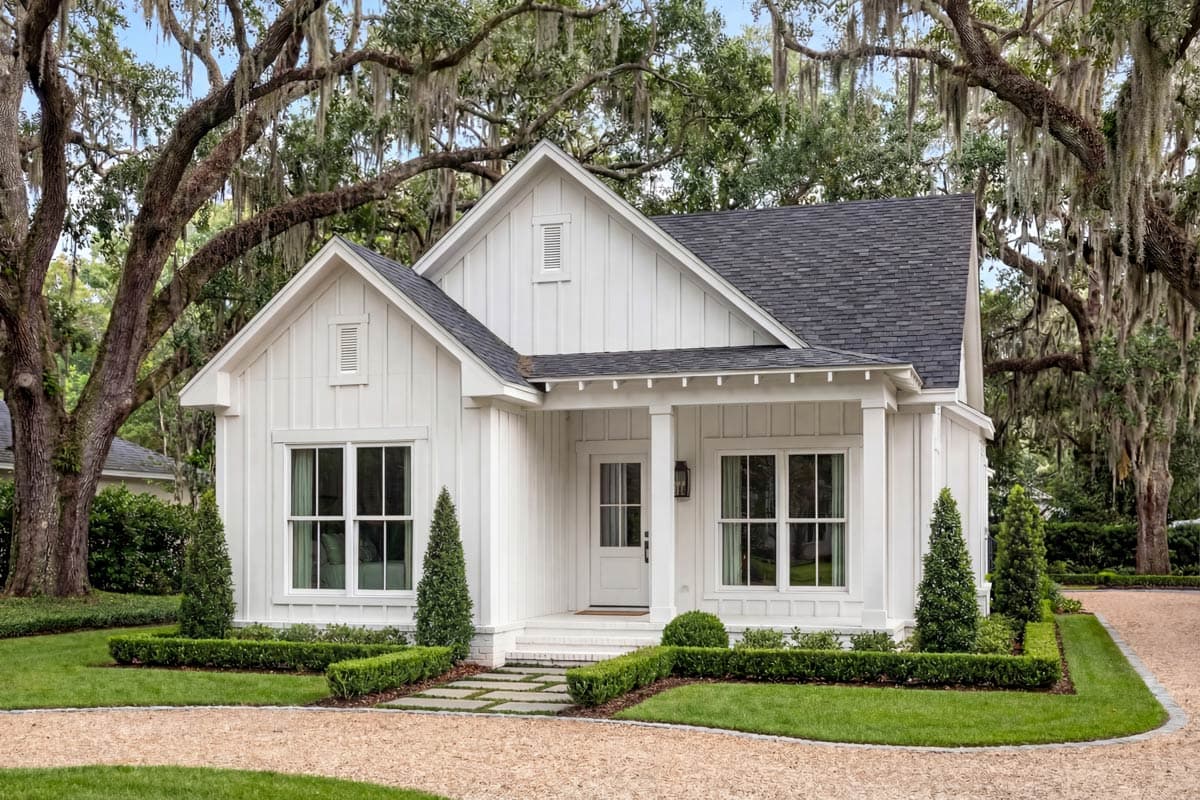 A white farmhouse with a dark roof and manicured landscaping sits beneath large oak trees draped with Spanish moss, gravel driveway.