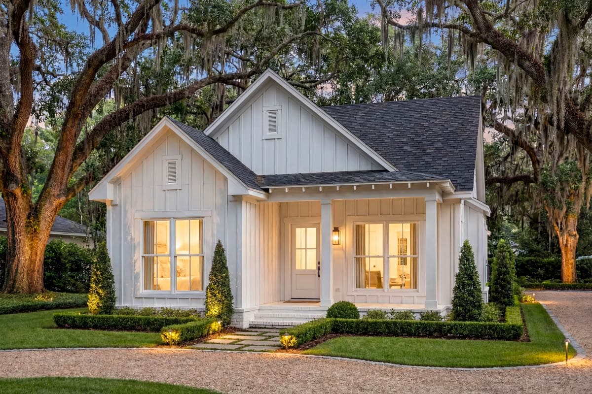 A charming white cottage with a porch, framed by mature oak trees. The warm light from the windows creates an inviting ambiance.