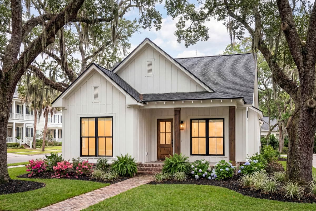A charming white house with black-framed windows, a brick path, and lush landscaping sits under large, moss-draped trees on a sunny day.