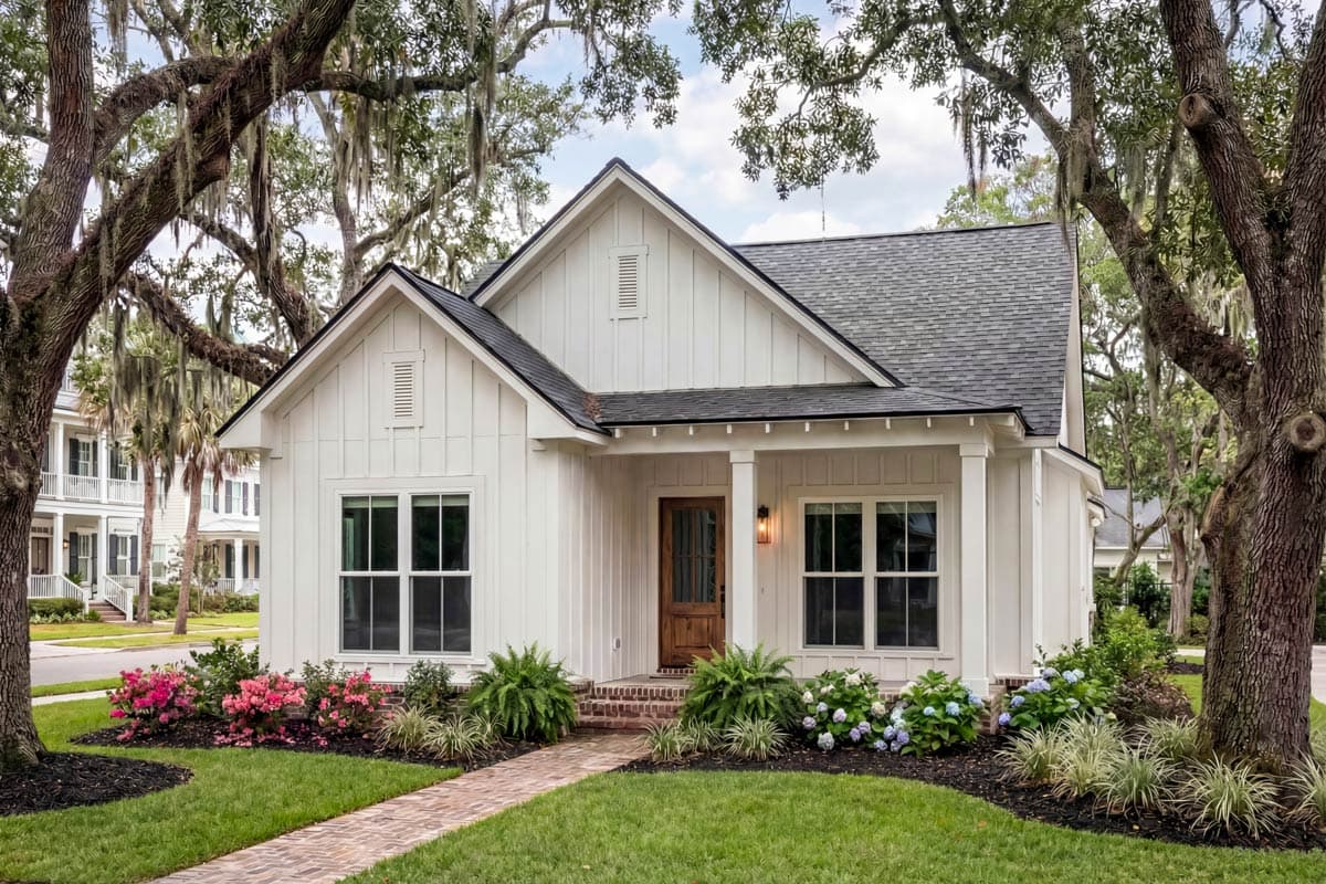 A white clapboard house with a dark roof is framed by large oak trees. A brick path leads to the front door, surrounded by landscaped gardens.