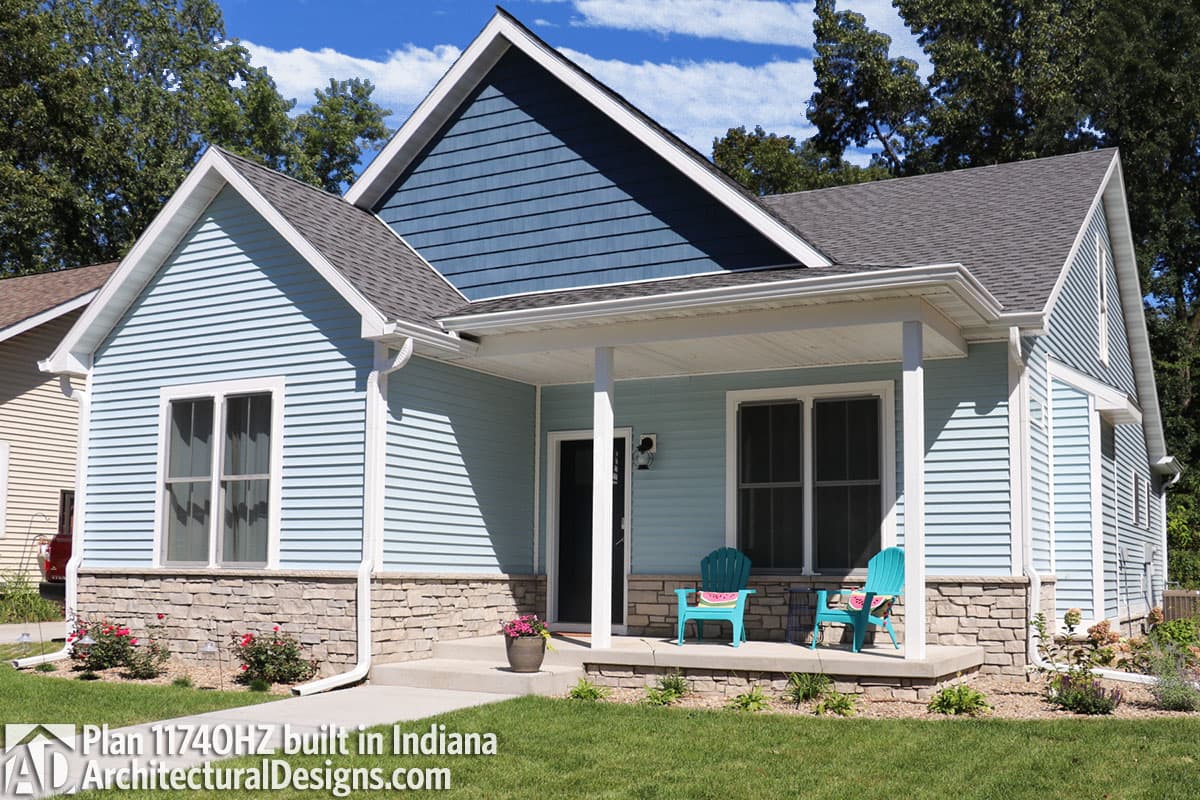 A light blue house with a covered porch and stone accents. Two teal chairs sit on the porch, and the front yard has lush green grass and flowerbeds.
