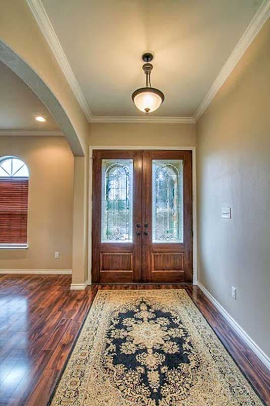 Interior view of an entryway with double wood doors featuring arched glass panels, a decorative rug, and hardwood floors.