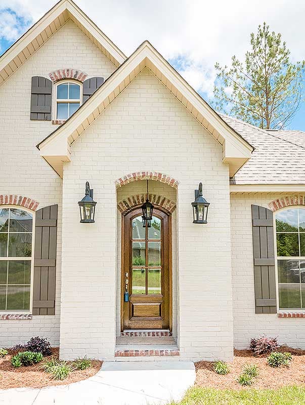 House plan exterior: white brick facade with arched wooden door, gabled roof, and dark louvered shutters on windows.