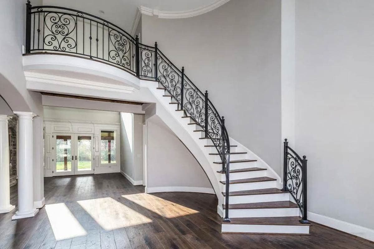 Interior view of curved staircase with ornate iron railing and white risers, leading to a balcony with matching railing. Tall white columns visible.