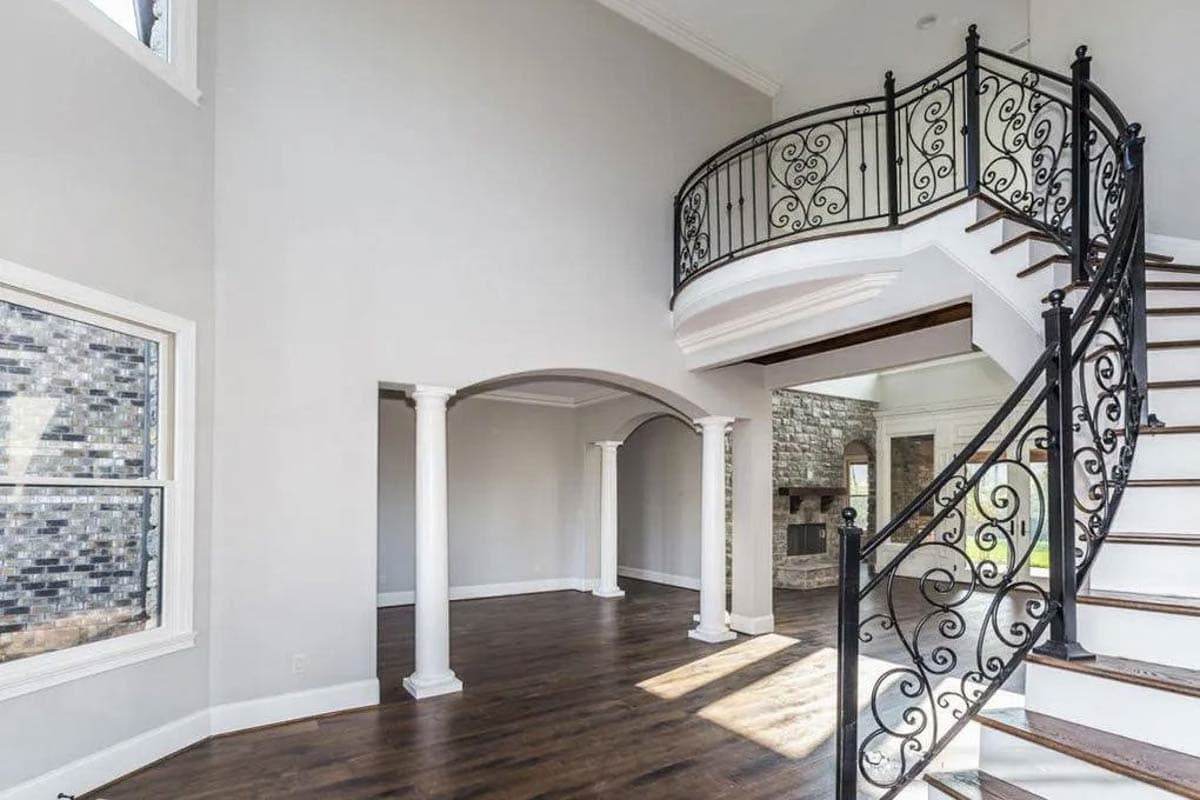 Interior view showing a grand foyer with a spiral staircase, decorative iron railings, columns, and arched doorways leading to a stone fireplace.