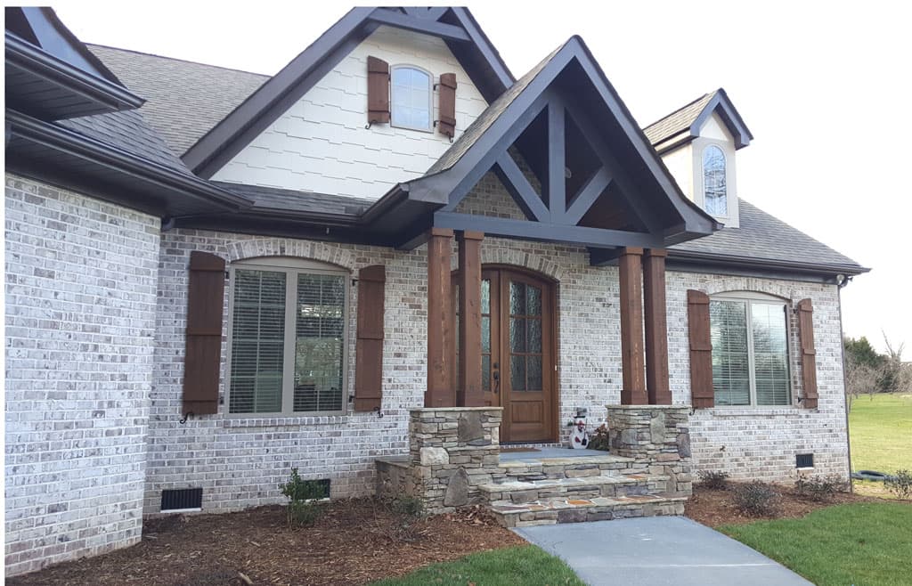 House plan exterior: Two-story dwelling with stone-clad foundation, brick exterior, gabled dormer, and timber-framed porch with arched double doors.