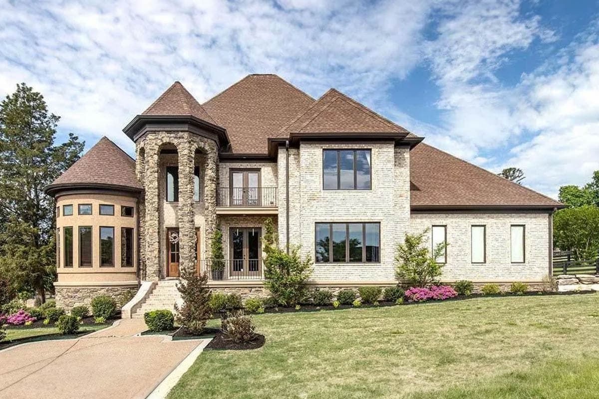 House plan exterior. Two-story with stone facade, turret, balcony, and multi-gabled roof. Entry features arched portico and stairs.