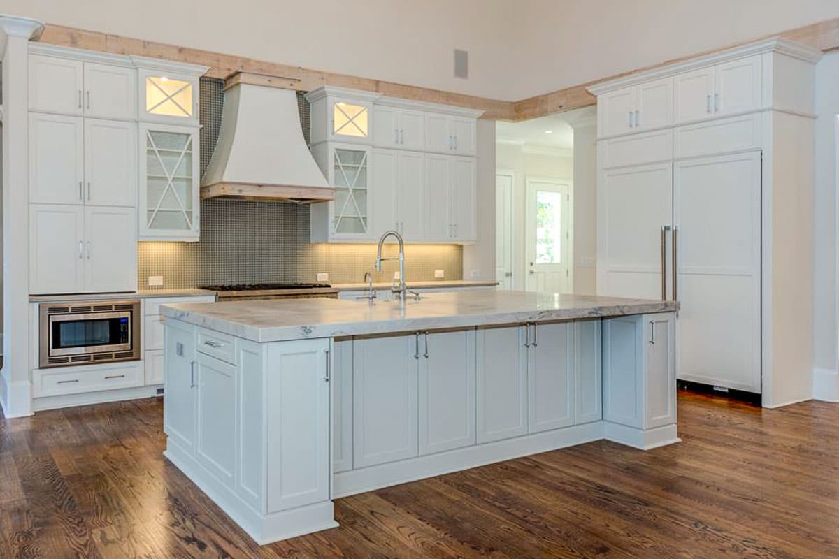 Interior view of a white kitchen featuring a large island with a marble countertop, custom cabinetry, a modern range hood, and built-in microwave.