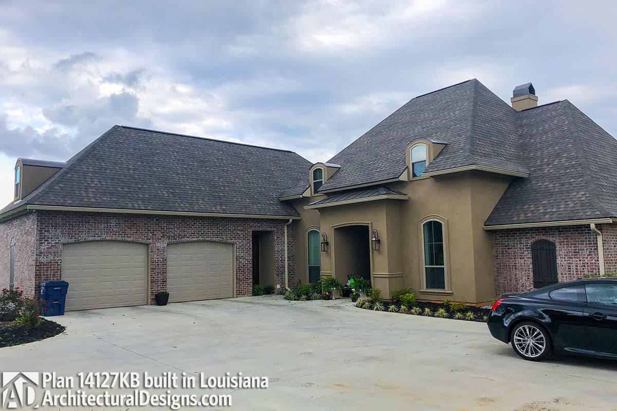 House plan exterior: Two-story home with a hip roof, stucco facade, brick accents, arched entryway, and a three-car garage.