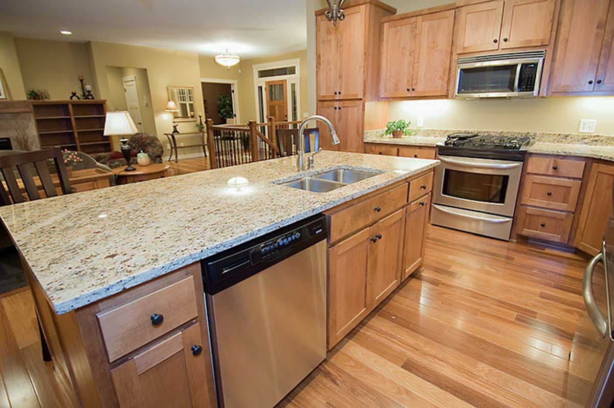 Kitchen interior with island sink, dishwasher, range, microwave, wooden cabinets, granite countertops, and adjoining living area.
