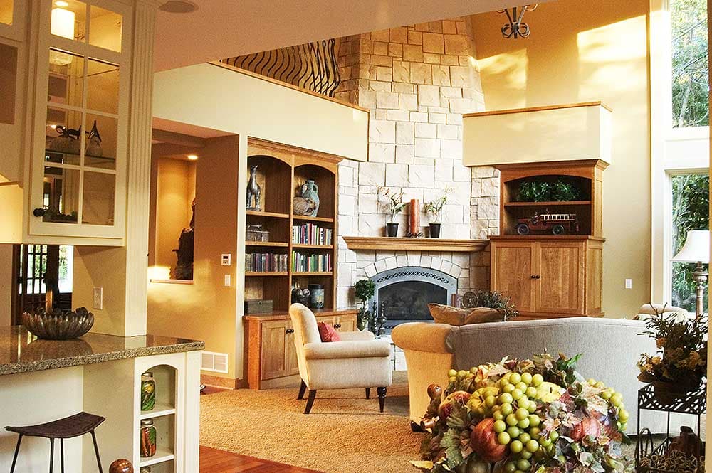 Interior view of a living room featuring a stone fireplace, built-in bookshelves, and a kitchen counter.