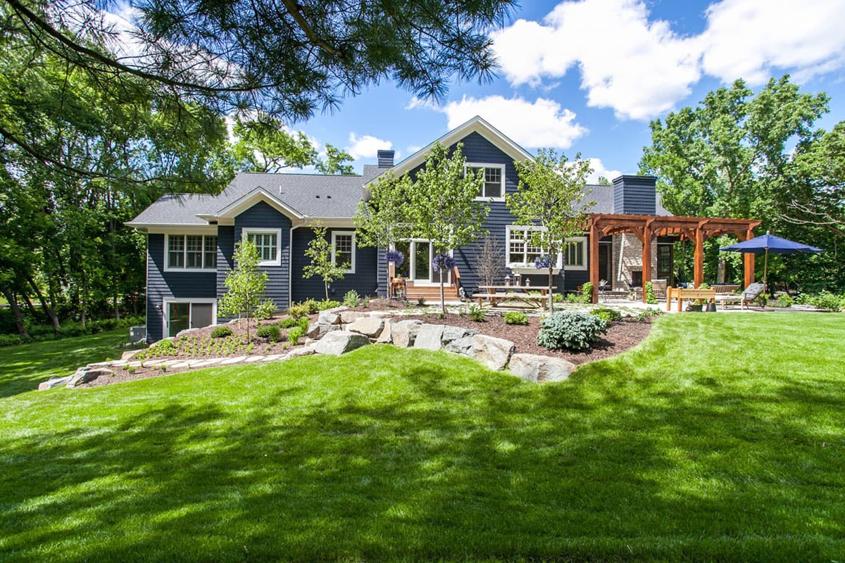 House plan exterior with dark blue siding, gable dormers, a prominent chimney, and a wooden pergola with an outdoor seating area.