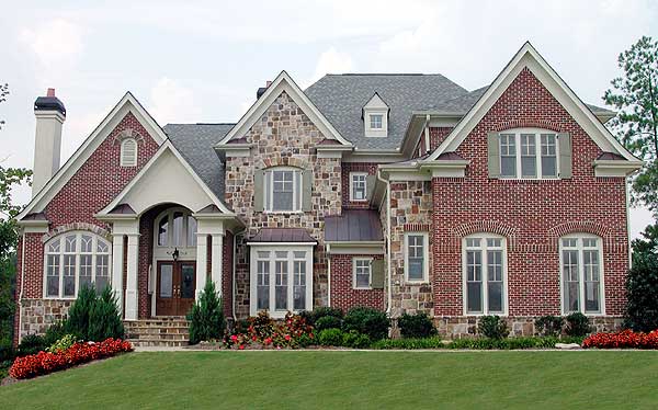 House plan exterior: Red brick two-story home with stone accents, prominent gables, arched entryway with columns, and a bay window.