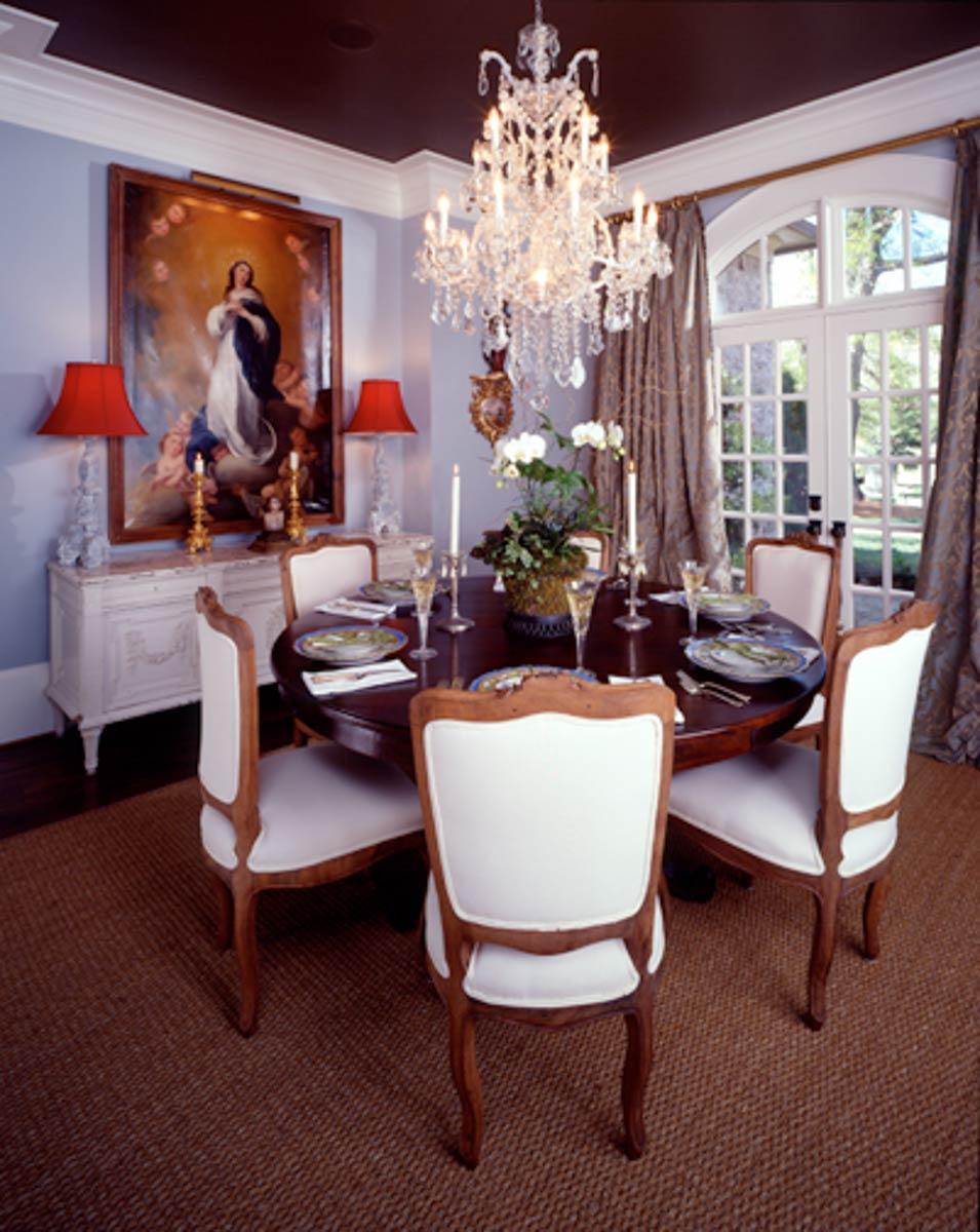 Dining room with round table set for six, ornate chandelier, large arched window, and traditional buffet with artwork.