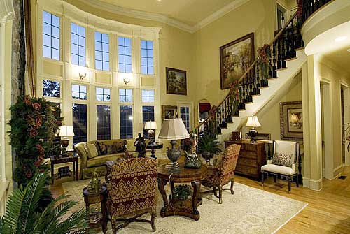 Interior view of a grand living room with a two-story curved window wall, ornate staircase, and plush seating.