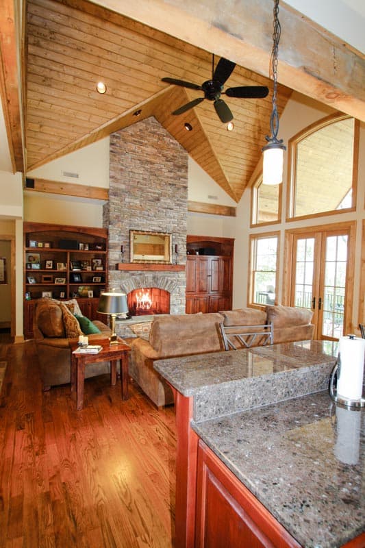 Great room interior with vaulted wood-beamed ceiling, stone fireplace, built-in bookshelves, and large windows.