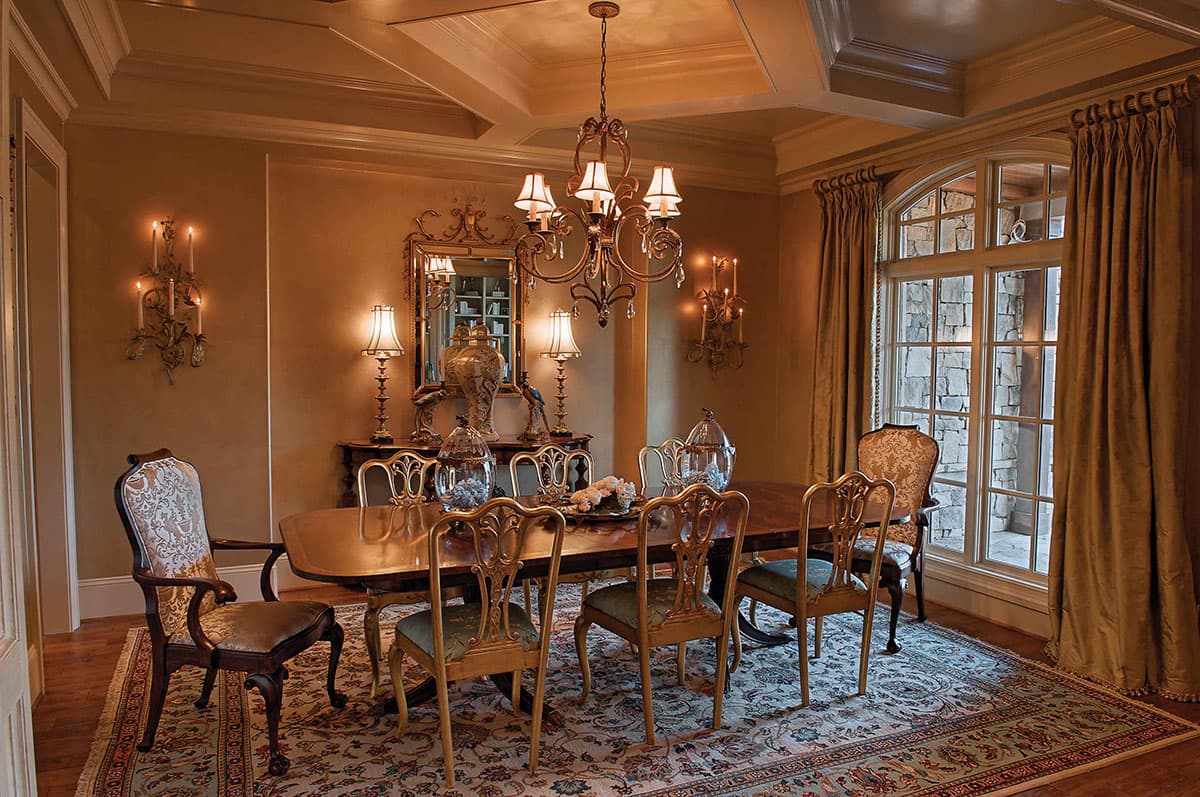 Interior view of a dining room with coffered ceiling, oval dining table, ornate chairs, and large mullioned window.
