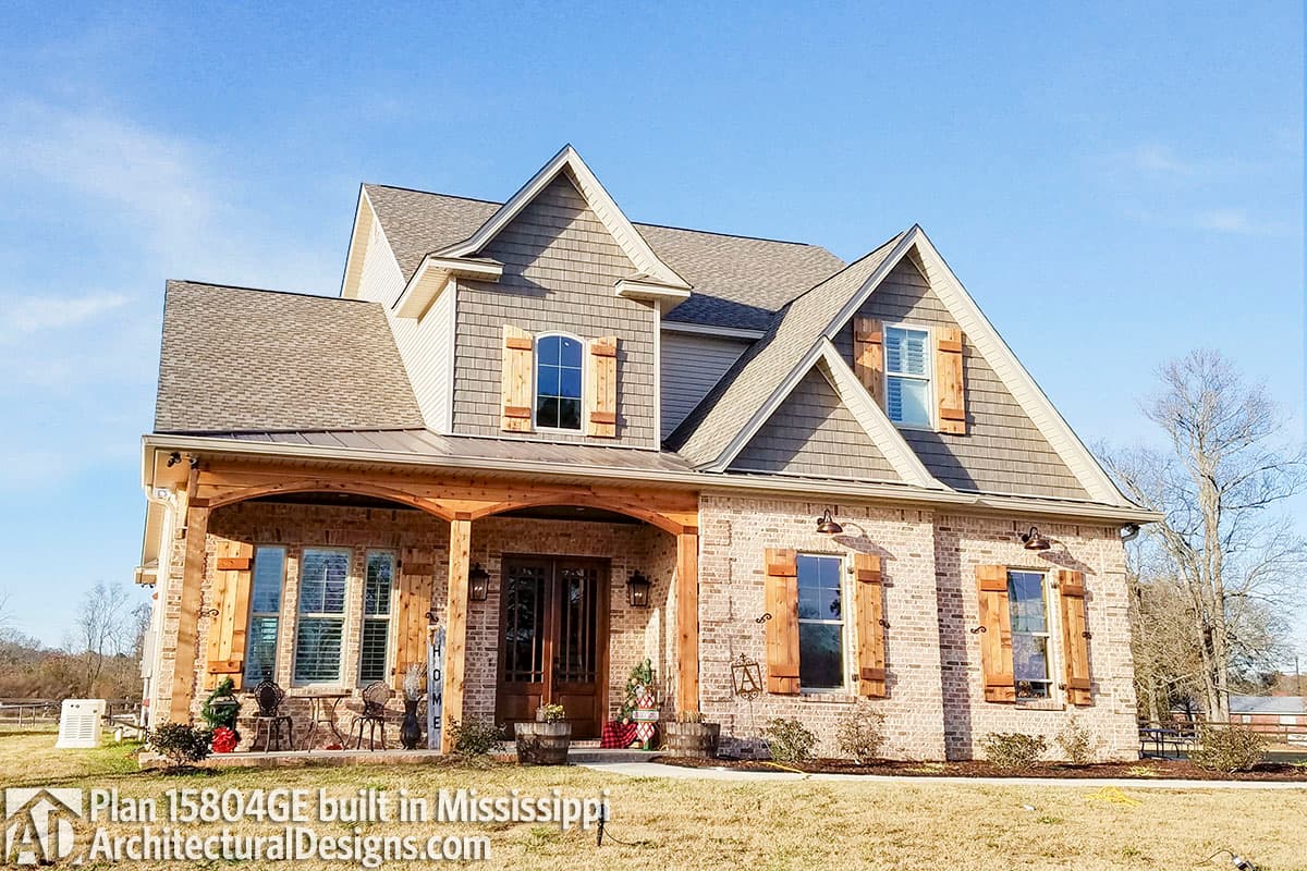 House plan exterior: Two-story Modern Farmhouse with brick facade, covered porch with wood beams, arched gable dormers, and cedar shutters.