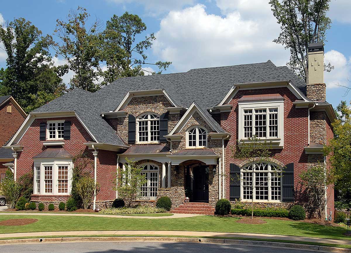 House plan exterior featuring brick and stone facade, multiple gables, arched windows with shutters, bay windows, and a prominent brick chimney.