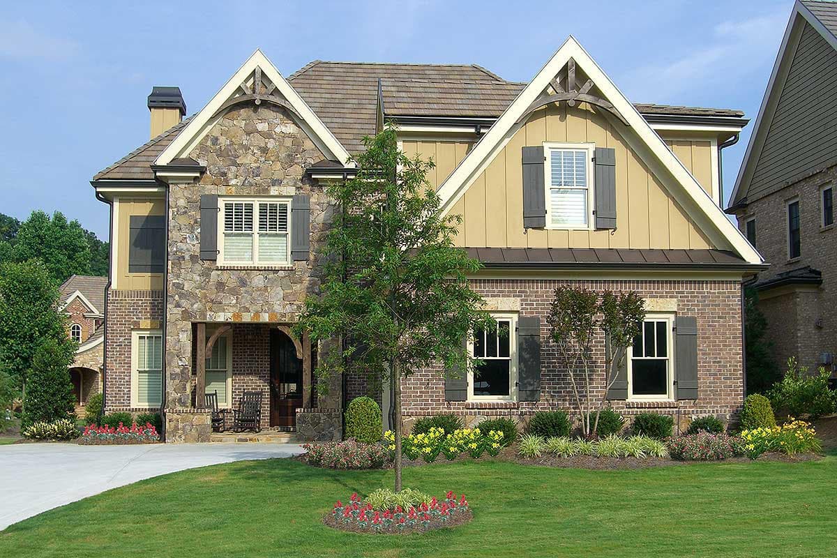 Two-story house plan exterior with stone and brick facade, prominent gables, arched entry porch, and multiple dormers.