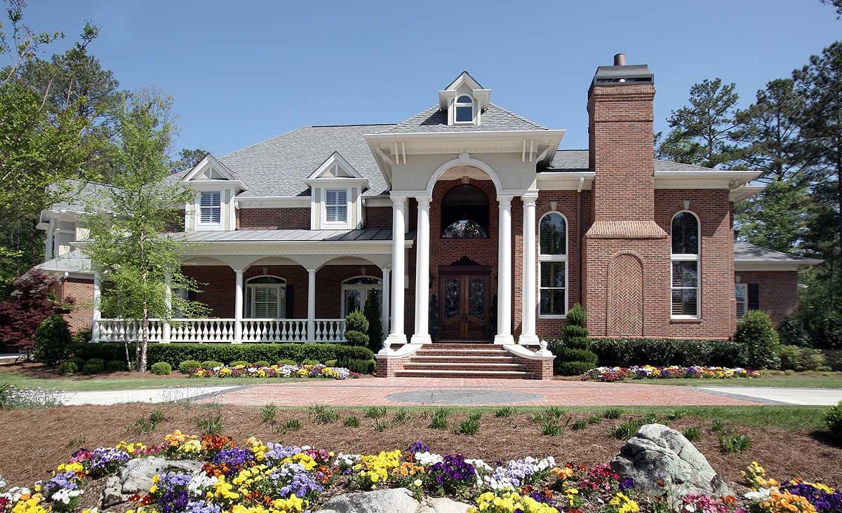 House plan exterior: Two-story brick home with wraparound porch, prominent portico, dual gables, and arched dormers.