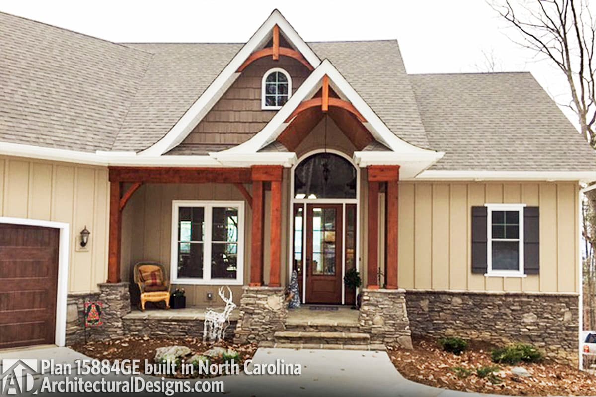 House plan exterior with a covered porch, stone accents, wood trim, arched entry, and a prominent gable with decorative rafter tails.