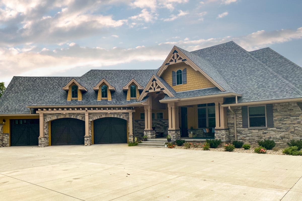 House plan exterior with stone veneer, gabled roof, and arched garage entry. Features include dormers and a covered porch.