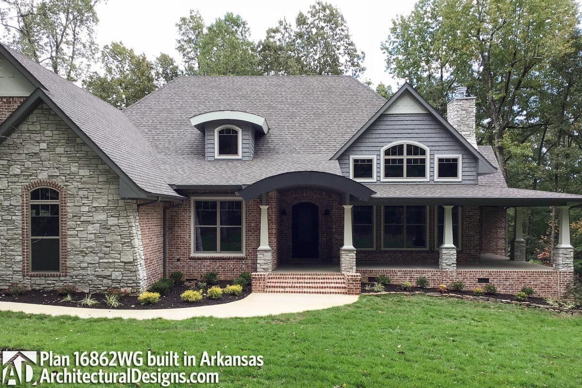 House plan exterior: Two-story home with stone and brick facade, arched dormer, covered front porch, and prominent stone chimney.