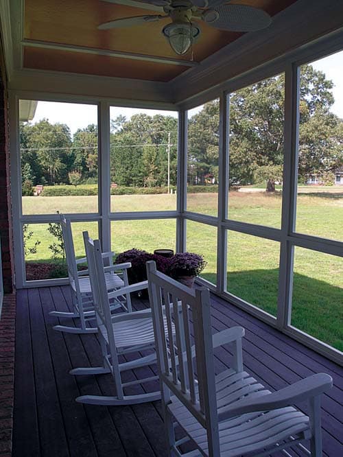 Screened porch with white rocking chairs, dark composite decking, and a ceiling fan, overlooking a grassy lawn and trees.