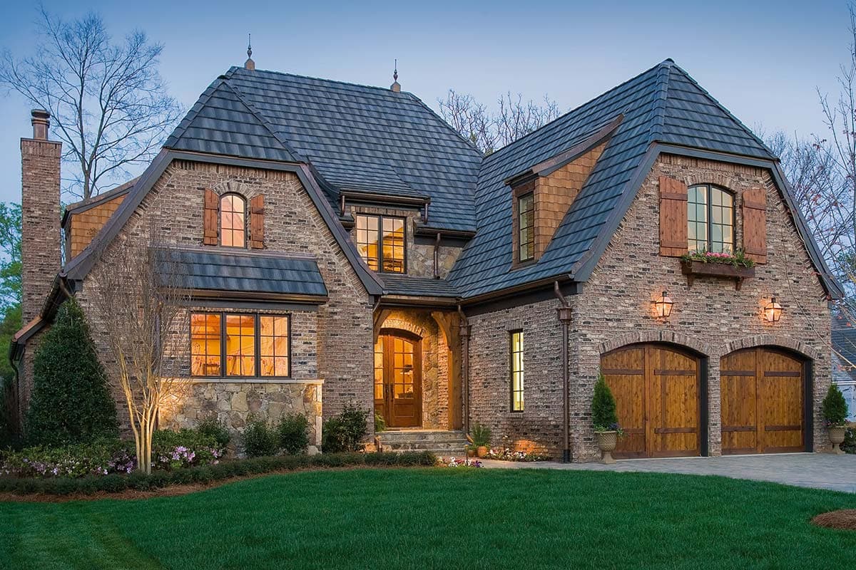 House plan exterior featuring a brick facade, steeply pitched rooflines, arched windows with wooden shutters, and a dual-car garage.