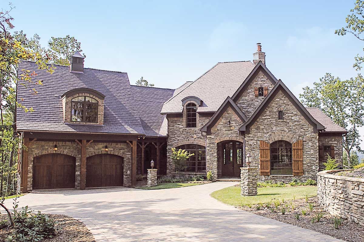 House plan exterior with stone facade, steep rooflines, arched windows, and a three-car side-entry garage.