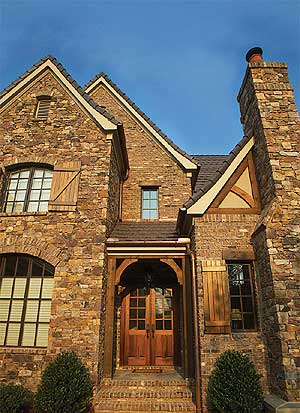 House plan exterior of a stone-clad home with gabled dormers, arched windows, wood shutters, and a covered entry with double doors.
