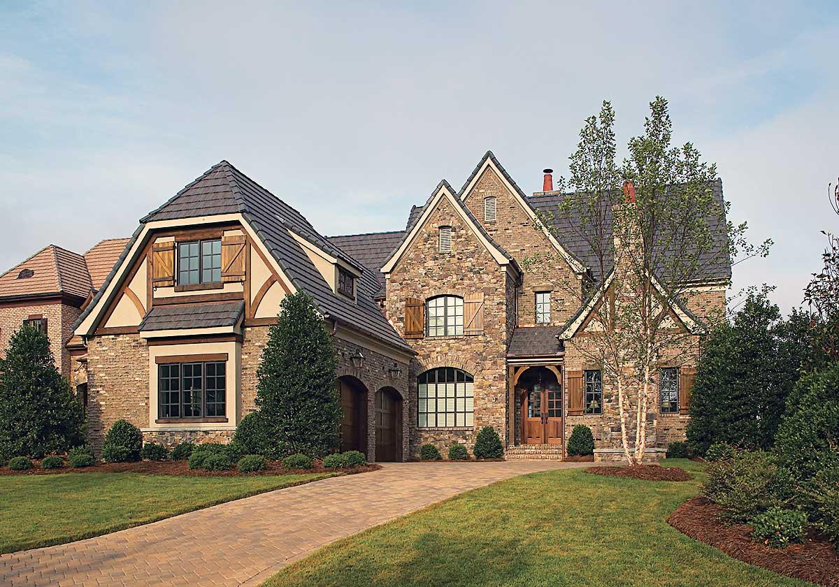 House plan exterior: Stone and stucco two-story home with a steeply pitched roof, arched windows, wood shutters, and a double garage.