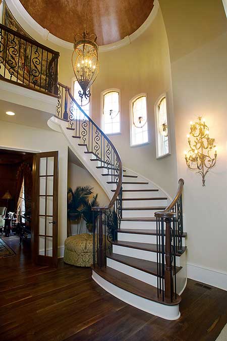 Curved staircase with wrought iron railing, arched windows, and ornate chandelier in a grand foyer.