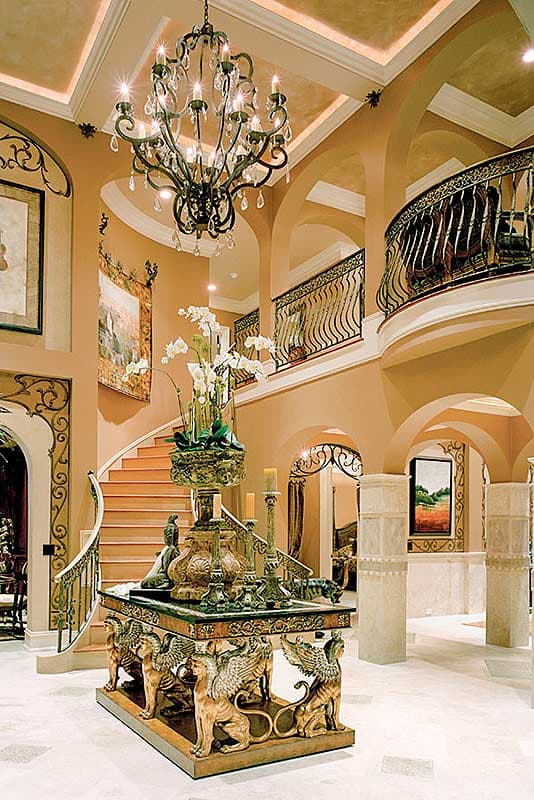 Grand foyer with a curved staircase, ornate chandelier, and sculptural pedestal table.