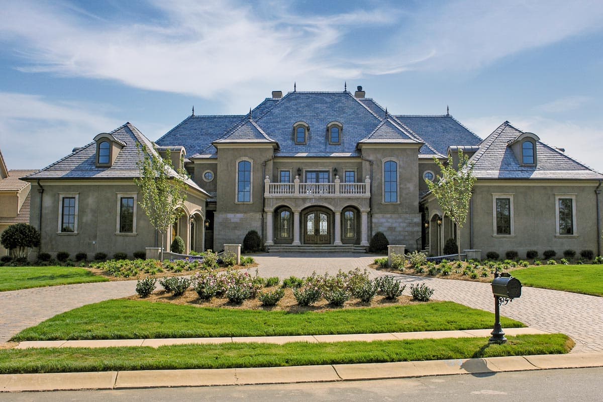 House plan exterior of large two-story home with mansard roof, arched windows, stone accents, and central balcony.