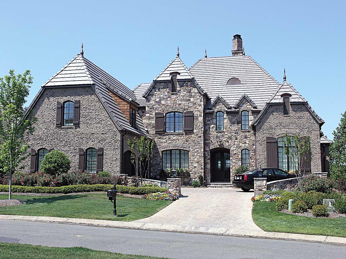House plan exterior: stone facade, multi-story structure with prominent gables, arched windows, and dark wood shutters.