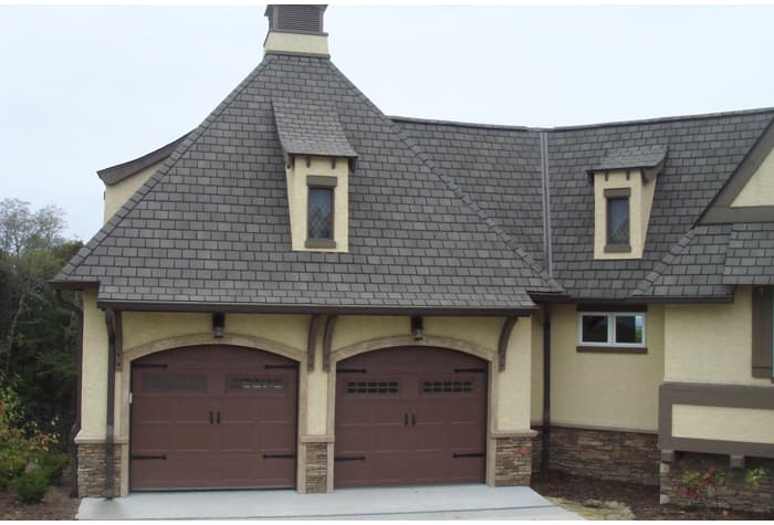 House plan exterior featuring a steeply pitched hip roof, two dormers, and a dual garage with arched wood doors.
