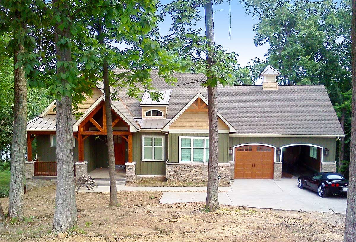 House plan exterior with exposed timber frame porch, stone base, dormer, and a two-car garage with arched doors.